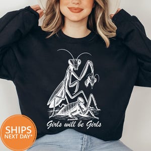 May include: A black long-sleeve sweatshirt featuring a white graphic illustration of a praying mantis holding another insect's head above a headless insect body. The text "Girls will be Girls" is printed in white script below the illustration.