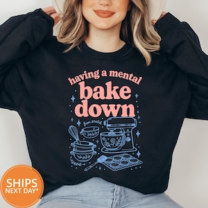May include: A black long-sleeve sweatshirt featuring the pink text "having a mental bake down" and the blue text "from scratch". The graphic includes line art of a stand mixer, mixing bowls, a whisk, and a tray of cookies with small star accents.