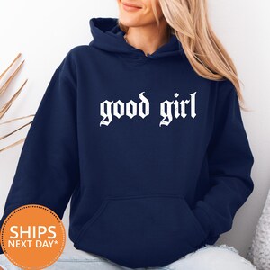 May include: Navy blue hoodie with the phrase "good girl" in white gothic font across the chest. The hoodie has a drawstring hood and a front pocket. The model is wearing the hoodie.