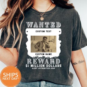 May include: Dark gray t-shirt with a sepia-toned image of a person and the text "WANTED" above and "REWARD $1 MILLION DOLLARS" below. The shirt includes space for custom text and name.