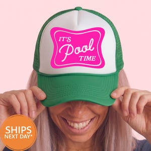 May include: A green and white trucker hat featuring a bright pink rectangular graphic with rounded corners. The graphic contains the white text IT'S Pool TIME in a mix of block and script fonts. The hat has a mesh back and a solid front panel.