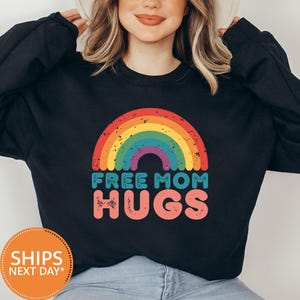 May include: Black sweatshirt featuring a rainbow graphic with the words "FREE MOM HUGS" in teal and pink. The design is centered on the chest. The sweatshirt is a casual, comfortable style.