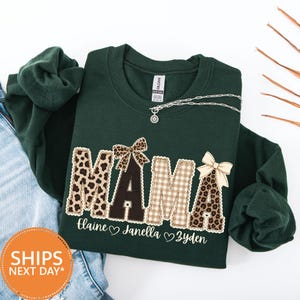 May include: A dark green long-sleeve sweatshirt featuring the word MAMA in large letters with leopard print and plaid patterns. The letters are decorated with small bows. Below the word, the names Elaine, Janella, and Zyden are printed.