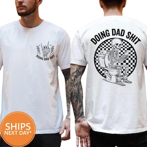May include: A white t-shirt featuring a black graphic on the front chest of a skeleton hand making a rock-and-roll gesture with the text DOING DAD SHIT. The back displays a skeleton sitting on a toilet using a phone with a checkered background.