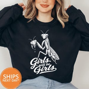May include: A black long-sleeve sweatshirt featuring a white graphic print of a praying mantis holding another insect head. Below the insect, the text reads "Girls will be Girls" in a bold, white, cursive-style font across the chest area.