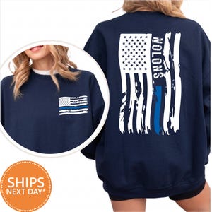 May include: A navy blue crewneck sweatshirt featuring a distressed American flag graphic on the back with a vertical blue stripe and the text NOLONS. A smaller version of the same distressed flag graphic is printed on the front left chest area.
