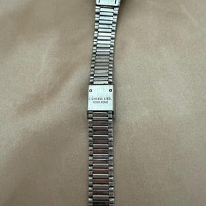 K&ouml;nnte beinhalten: Eine silberfarbene Armbanduhr mit einem Armband aus Edelstahl. Das Armband besteht aus kleinen, rechteckigen Gliedern. Der Verschluss ist mit "STAINLESS STEEL HONG KONG" gekennzeichnet. Das Zifferblatt der Uhr ist oben sichtbar.