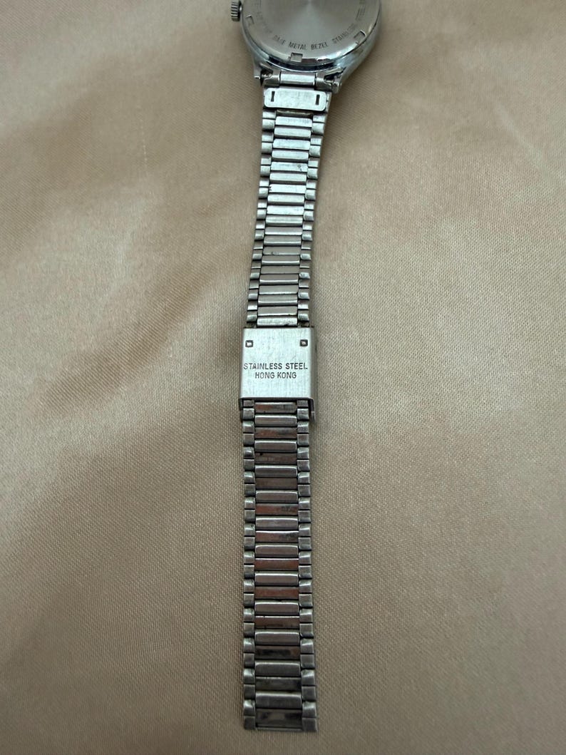 K&ouml;nnte beinhalten: Eine silberfarbene Armbanduhr mit einem Armband aus Edelstahl. Das Armband besteht aus kleinen, rechteckigen Gliedern. Der Verschluss ist mit "STAINLESS STEEL HONG KONG" gekennzeichnet. Das Zifferblatt der Uhr ist oben sichtbar.