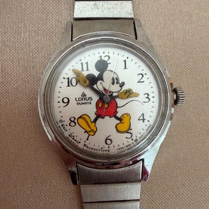 K&ouml;nnte beinhalten: Eine silberfarbene Lorus-Quarzuhr mit wei&szlig;em Zifferblatt, auf dem Mickey Mouse in roter Hose, gelben Schuhen und schwarzen Handschuhen abgebildet ist. Die Uhr hat ein silbernes Armband und ein kleines Zifferblatt an der Seite.