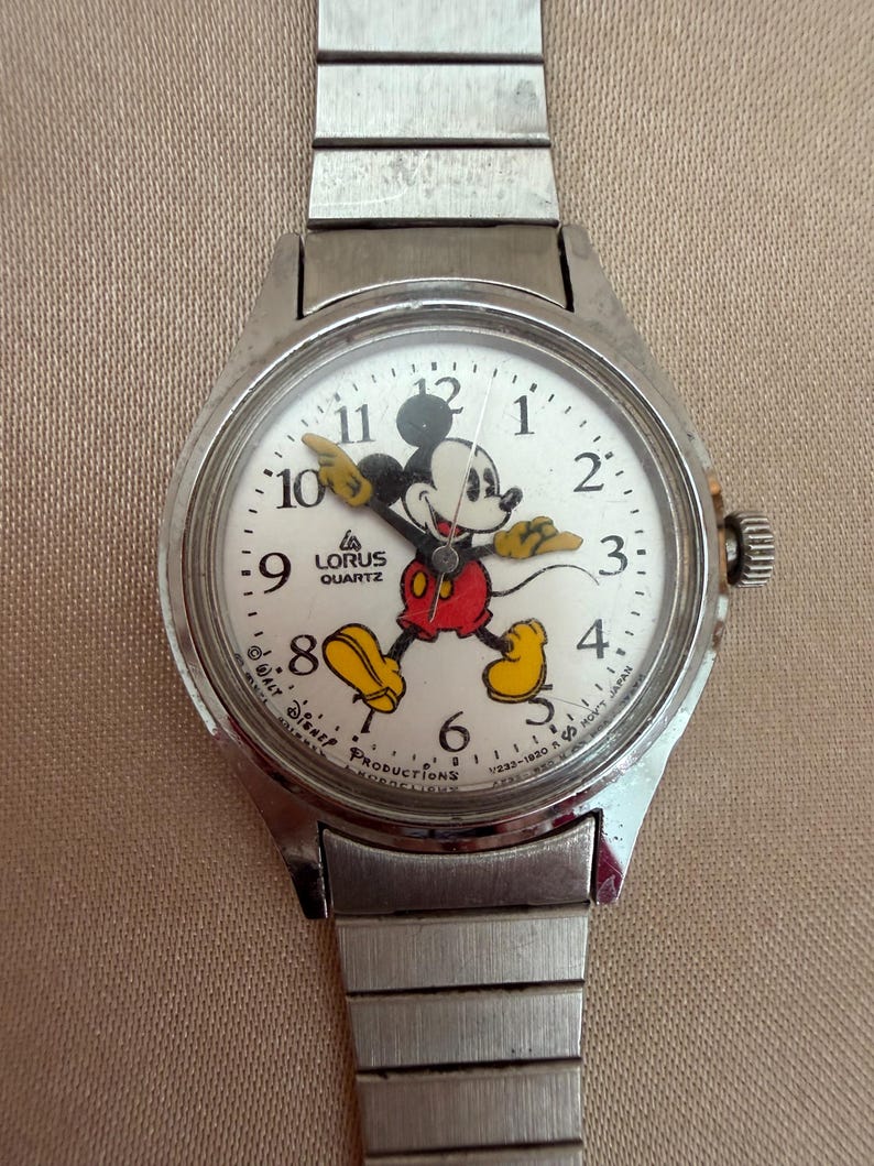 K&ouml;nnte beinhalten: Eine silberfarbene Lorus-Quarzuhr mit wei&szlig;em Zifferblatt, auf dem Mickey Mouse in roter Hose, gelben Schuhen und schwarzen Handschuhen abgebildet ist. Die Uhr hat ein silbernes Armband und ein kleines Zifferblatt an der Seite.
