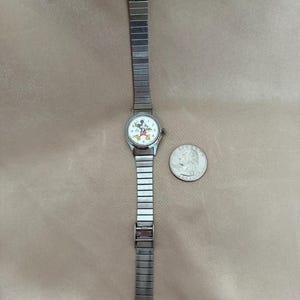 K&ouml;nnte beinhalten: Eine silberfarbene Armbanduhr mit wei&szlig;em Zifferblatt mit Mickey Mouse. Die Uhr hat ein rechteckiges Gliederarmband und einen Verschluss. Eine M&uuml;nze liegt neben der Uhr zum Gr&ouml;&szlig;envergleich.