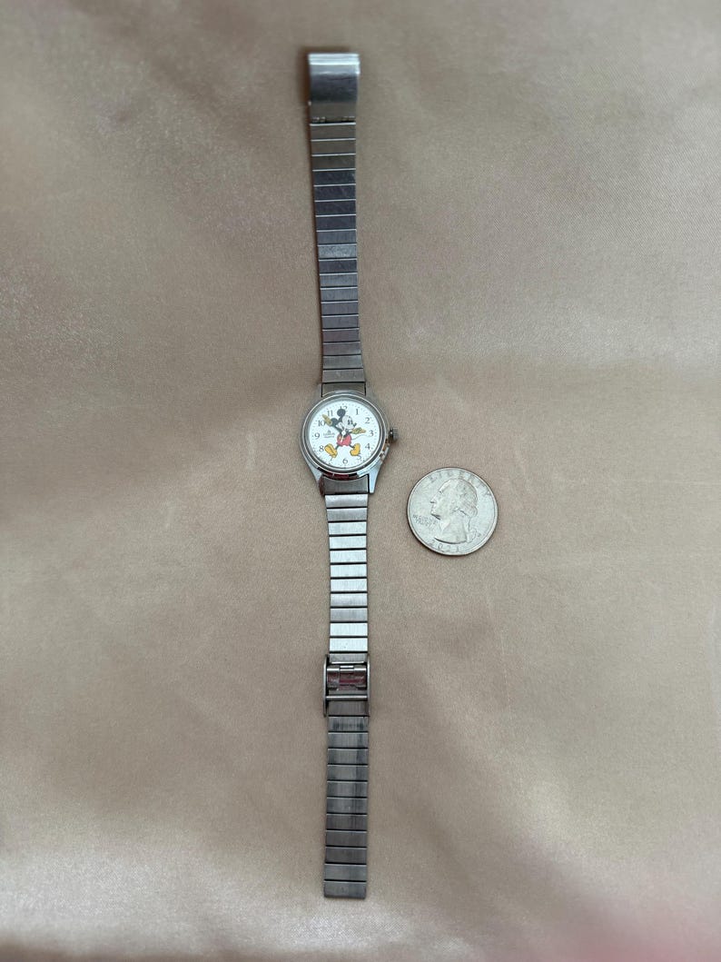 K&ouml;nnte beinhalten: Eine silberfarbene Armbanduhr mit wei&szlig;em Zifferblatt mit Mickey Mouse. Die Uhr hat ein rechteckiges Gliederarmband und einen Verschluss. Eine M&uuml;nze liegt neben der Uhr zum Gr&ouml;&szlig;envergleich.