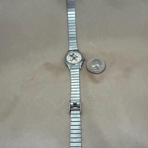 K&ouml;nnte beinhalten: Silberfarbene Armbanduhr mit Mickey Mouse-Design auf dem Zifferblatt. Die Uhr hat ein rechteckiges, dehnbares Armband. Ein M&uuml;nze liegt neben der Uhr zum Gr&ouml;&szlig;envergleich.
