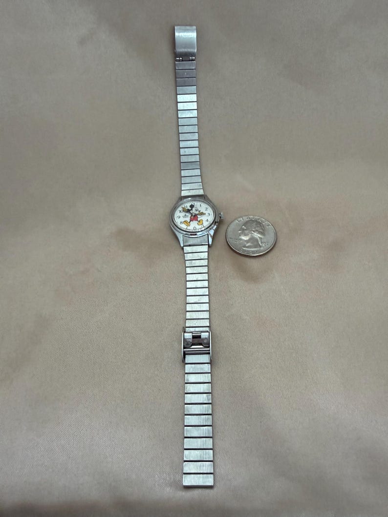 K&ouml;nnte beinhalten: Silberfarbene Armbanduhr mit Mickey Mouse-Design auf dem Zifferblatt. Die Uhr hat ein rechteckiges, dehnbares Armband. Ein M&uuml;nze liegt neben der Uhr zum Gr&ouml;&szlig;envergleich.