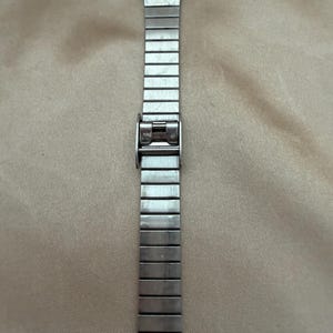 K&ouml;nnte beinhalten: Eine silberfarbene Armbanduhr mit rechteckigem Gliederarmband. Das Zifferblatt der Uhr ist sichtbar, mit hellem Hintergrund und schwarzen Ziffern und Zeigern. Das Armband besteht aus verbundenen Metallsegmenten.