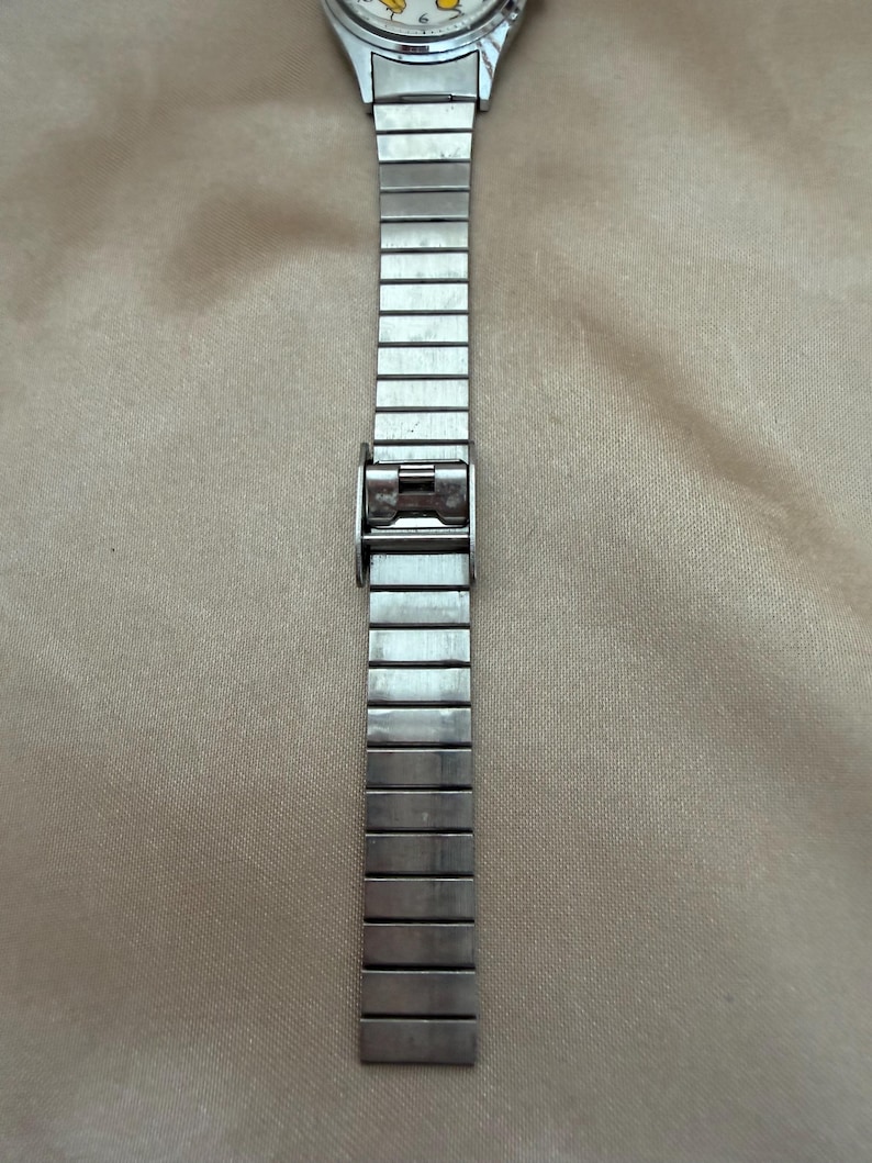 K&ouml;nnte beinhalten: Eine silberfarbene Armbanduhr mit rechteckigem Gliederarmband. Das Zifferblatt der Uhr ist sichtbar, mit hellem Hintergrund und schwarzen Ziffern und Zeigern. Das Armband besteht aus verbundenen Metallsegmenten.