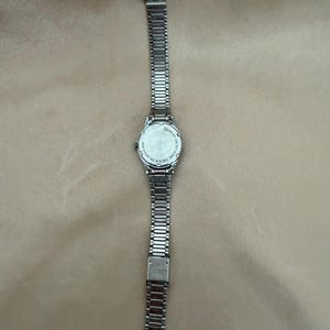 K&ouml;nnte beinhalten: Eine silberfarbene Armbanduhr mit rechteckigem Zifferblatt und einem Metallarmband mit Gliedern. Die Uhr wird auf einer neutralfarbenen Oberfl&auml;che pr&auml;sentiert. Das Armband ist etwa 2 cm breit.
