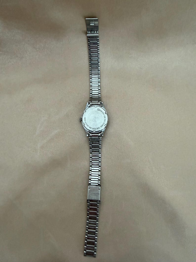 K&ouml;nnte beinhalten: Eine silberfarbene Armbanduhr mit rechteckigem Zifferblatt und einem Metallarmband mit Gliedern. Die Uhr wird auf einer neutralfarbenen Oberfl&auml;che pr&auml;sentiert. Das Armband ist etwa 2 cm breit.