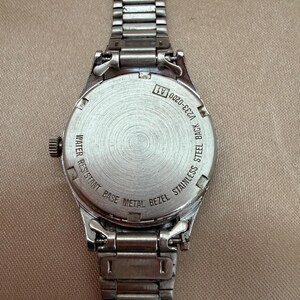 K&ouml;nnte beinhalten: Eine silberfarbene Armbanduhr mit Edelstahlr&uuml;cken und einer L&uuml;nette aus unedlem Metall. Die Uhr hat ein Metallarmband und ein rundes Zifferblatt. Auf der R&uuml;ckseite der Uhr steht "WATER RESISTANT BASE METAL BEZEL STAINLESS STEEL BACK V233-0200 A1".