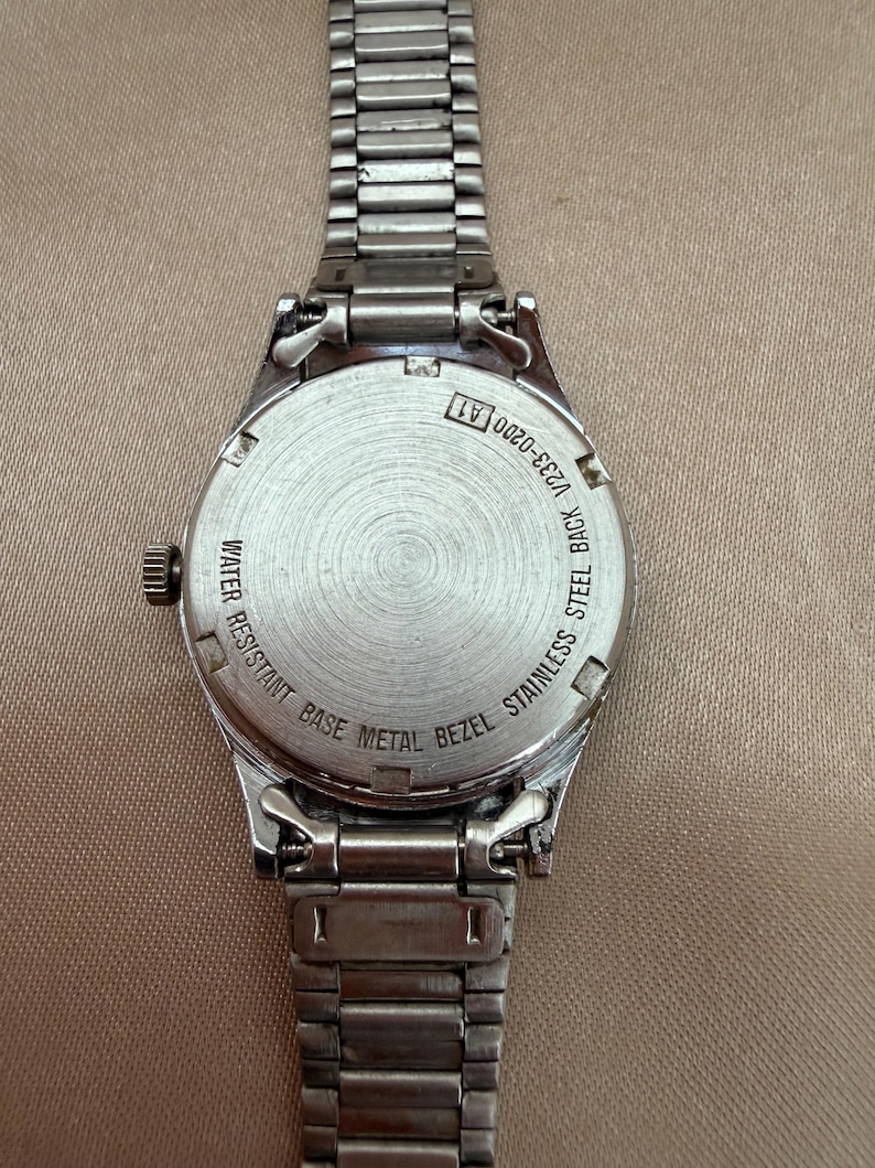 K&ouml;nnte beinhalten: Eine silberfarbene Armbanduhr mit Edelstahlr&uuml;cken und einer L&uuml;nette aus unedlem Metall. Die Uhr hat ein Metallarmband und ein rundes Zifferblatt. Auf der R&uuml;ckseite der Uhr steht "WATER RESISTANT BASE METAL BEZEL STAINLESS STEEL BACK V233-0200 A1".