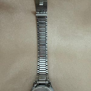 K&ouml;nnte beinhalten: Eine silberfarbene Uhr mit Metallarmband. Das Zifferblatt der Uhr ist rund mit silbernem R&uuml;cken. Das Armband besteht aus kleinen, rechteckigen Metallgliedern. Der Verschluss ist ein einfaches, rechteckiges Design. Die Uhr liegt auf einer beigen Oberfl&auml;che.