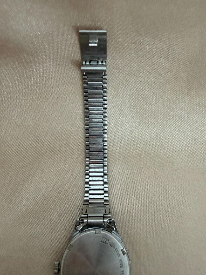 K&ouml;nnte beinhalten: Eine silberfarbene Uhr mit Metallarmband. Das Zifferblatt der Uhr ist rund mit silbernem R&uuml;cken. Das Armband besteht aus kleinen, rechteckigen Metallgliedern. Der Verschluss ist ein einfaches, rechteckiges Design. Die Uhr liegt auf einer beigen Oberfl&auml;che.