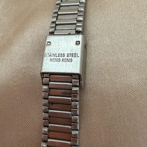 K&ouml;nnte beinhalten: Nahaufnahme eines silbernen Edelstahl-Uhrenarmbands. Das Armband hat ein rechteckiges Design. Auf dem Verschluss ist der Text "STAINLESS STEEL HONG KONG" zu sehen.