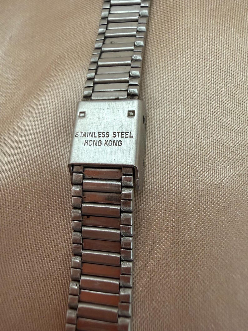 K&ouml;nnte beinhalten: Nahaufnahme eines silbernen Edelstahl-Uhrenarmbands. Das Armband hat ein rechteckiges Design. Auf dem Verschluss ist der Text "STAINLESS STEEL HONG KONG" zu sehen.