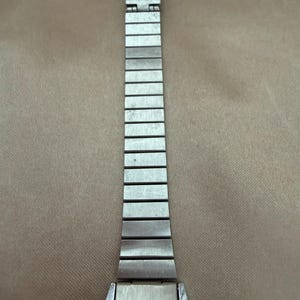K&ouml;nnte beinhalten: Eine silberfarbene Armbanduhr mit rechteckigem Zifferblatt und silbernem Metallarmband. Das Zifferblatt hat schwarze Ziffern und Zeiger auf wei&szlig;em Hintergrund. Das Armband besteht aus kleinen, verbundenen rechteckigen Gliedern.