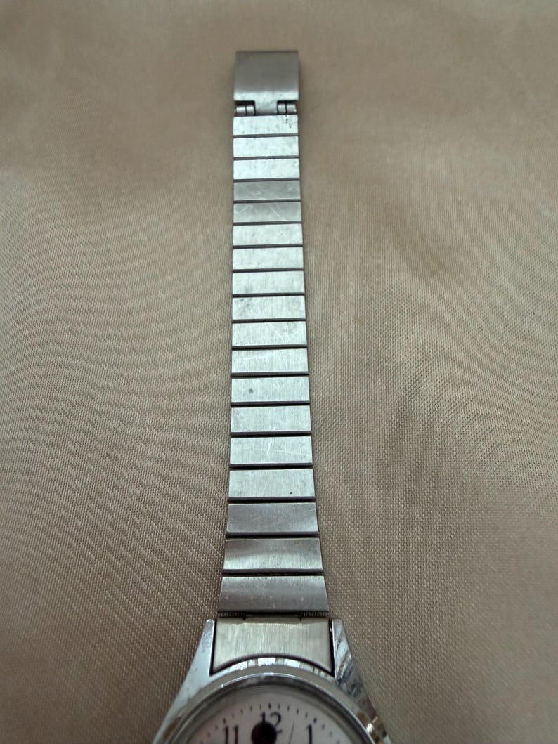 K&ouml;nnte beinhalten: Eine silberfarbene Armbanduhr mit rechteckigem Zifferblatt und silbernem Metallarmband. Das Zifferblatt hat schwarze Ziffern und Zeiger auf wei&szlig;em Hintergrund. Das Armband besteht aus kleinen, verbundenen rechteckigen Gliedern.