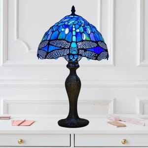 May include: A Tiffany-style table lamp with a blue dragonfly design. The lamp features a stained glass shade with a dragonfly motif in shades of blue and green. The base is dark brown with a decorative, ornate design.