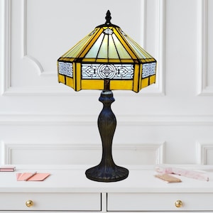 Könnte beinhalten: Eine Tischlampe aus Buntglas mit einem gelb-weißen geometrischen Schirm. Die Lampe hat einen dunkelbronzenen Fuß und Stiel mit floralem Design. Der Schirm hat eine vielseitige Form mit dekorativem Rand.
