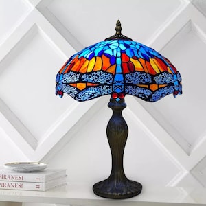 Könnte beinhalten: Eine Tischlampe aus Tiffany-Glas mit einem Libellenmotiv. Der Lampenschirm ist blau, orange und rot. Der Sockel ist dunkelbronzefarben.