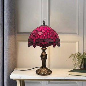 May include: A table lamp featuring a dome-shaped stained glass shade in vibrant pink with dark dragonfly motifs and embedded glass jewels. The lamp has a dark bronze-coloured metal base with a decorative, curved design and a black power cord.