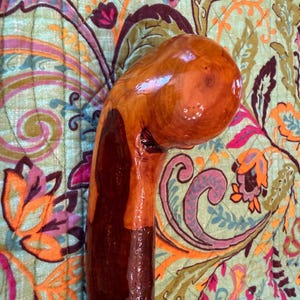 Puede incluir: Un bastón de madera hecho a mano con un acabado brillante. El bastón presenta un mango redondeado y un eje marrón oscuro, que pasa a un color marrón rojizo más claro cerca del mango. La veta de la madera es visible.