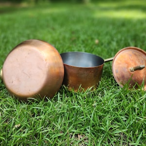 May include: A copper pot with a metallic interior and a matching circular lid. The pot features a long handle extending to the left, while the lid rests to the right. The exterior surface of the copper cookware displays a warm, polished finish.