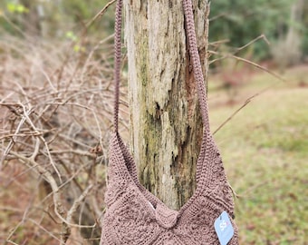 Könnte beinhalten: Eine handgefertigte, braune Häkeltasche mit Schulterriemen und langem Gurt, die an einem verwitterten Baumstumpf hängt. Die Tasche hat ein geometrisches Muster und einen Knopfverschluss. Ein kleines blaues Etikett ist an der Tasche befestigt.