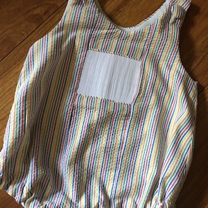 May include: A multi-colored striped baby romper with a white pocket and buttons. The romper is made of a soft, lightweight fabric and has a gathered bottom.
