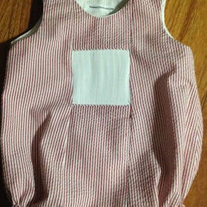 May include: A red and white striped baby onesie with a white patch on the front. The onesie has a button closure at the shoulders and a ruffled hem. The text "TreasuredMoments.com" is printed on the white patch.