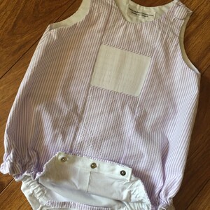 May include: A purple and white striped baby onesie with a white pocket and button closures. The onesie has a ruffled hem and is made of a soft, lightweight fabric. "Treasured Heirlooms.com Baby-So-Smick" is printed on the onesie.