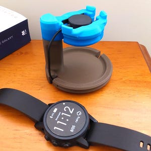 Puede incluir: Un reloj inteligente negro con esfera redonda que muestra las 11:12 y la fecha 05/08. En el fondo hay un soporte de carga azul y marrón. El soporte tiene una base circular y una cuna para el reloj.