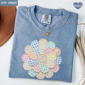 以下が含まれることがあります： 大きなカラフルな花のアップリケが付いた、水色のComfort Colors Tシャツ。 花びらは、ギンガムや花柄など、さまざまなパステル模様が特徴です。 ペンダント付きのゴールドネックレスがシャツの上にあります。