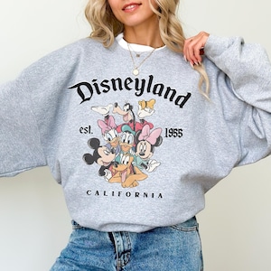 May include: A grey sweatshirt with the word "Disneyland" in black script, the text "est. 1955" and "CALIFORNIA". The sweatshirt features a graphic of Disney characters including Mickey Mouse, Minnie Mouse, Donald Duck, and Goofy.