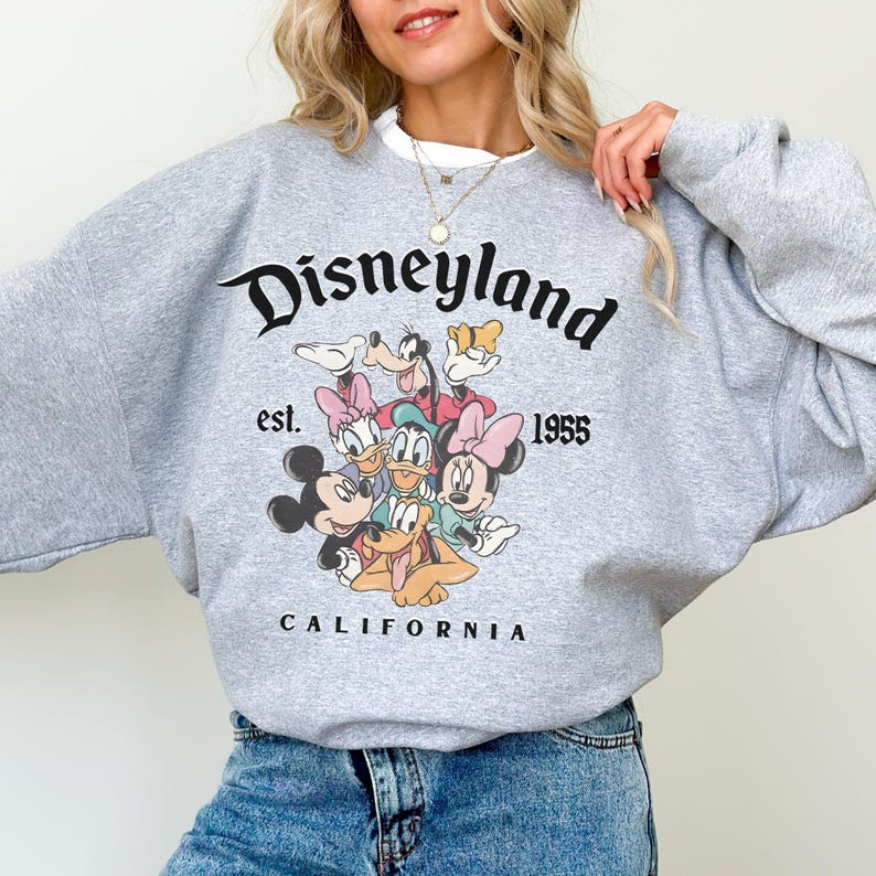 May include: A grey sweatshirt with the word "Disneyland" in black script, the text "est. 1955" and "CALIFORNIA". The sweatshirt features a graphic of Disney characters including Mickey Mouse, Minnie Mouse, Donald Duck, and Goofy.
