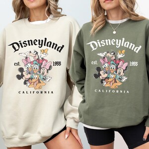 May include: Two sweatshirts, one beige and one olive green, with the Disneyland logo and cartoon characters including Mickey and Minnie Mouse. The text "Disneyland est. 1955 CALIFORNIA" is printed above the characters.