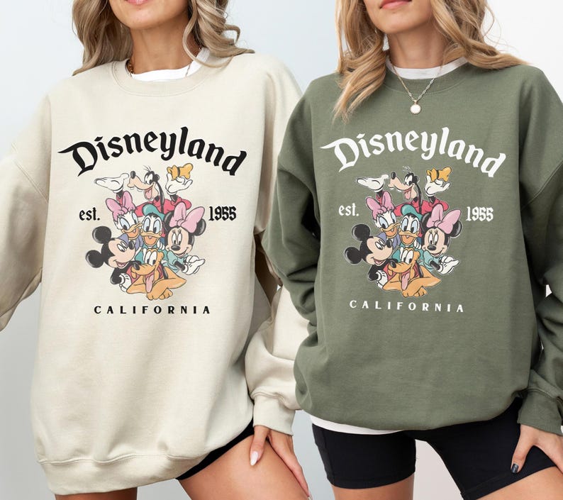 May include: Two sweatshirts, one beige and one olive green, with the Disneyland logo and cartoon characters including Mickey and Minnie Mouse. The text "Disneyland est. 1955 CALIFORNIA" is printed above the characters.