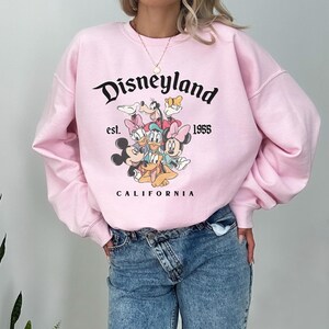 May include: A pale pink Disneyland sweatshirt with a graphic of Disney characters including Mickey and Minnie Mouse. The text "Disneyland est. 1955 California" is printed above and below the graphic in black.