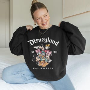 May include: Black sweatshirt featuring the Disneyland logo and a graphic of Mickey Mouse, Minnie Mouse, Donald Duck, Daisy Duck, Goofy, and Pluto. The text "Disneyland est. 1955 California" is printed on the front. The sweatshirt is a crew neck style.