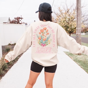 Puede incluir: Una sudadera beige con un diseño floral y el texto "Let all that you do be done in love 1 Corinthians 16:14".