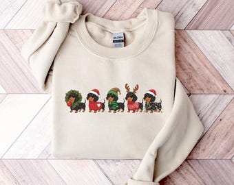 May include: A beige sweatshirt featuring a festive dachshund design. Five dachshunds are dressed in holiday attire, including Santa hats, reindeer antlers, and Christmas lights. The sweatshirt is folded on a wooden surface.